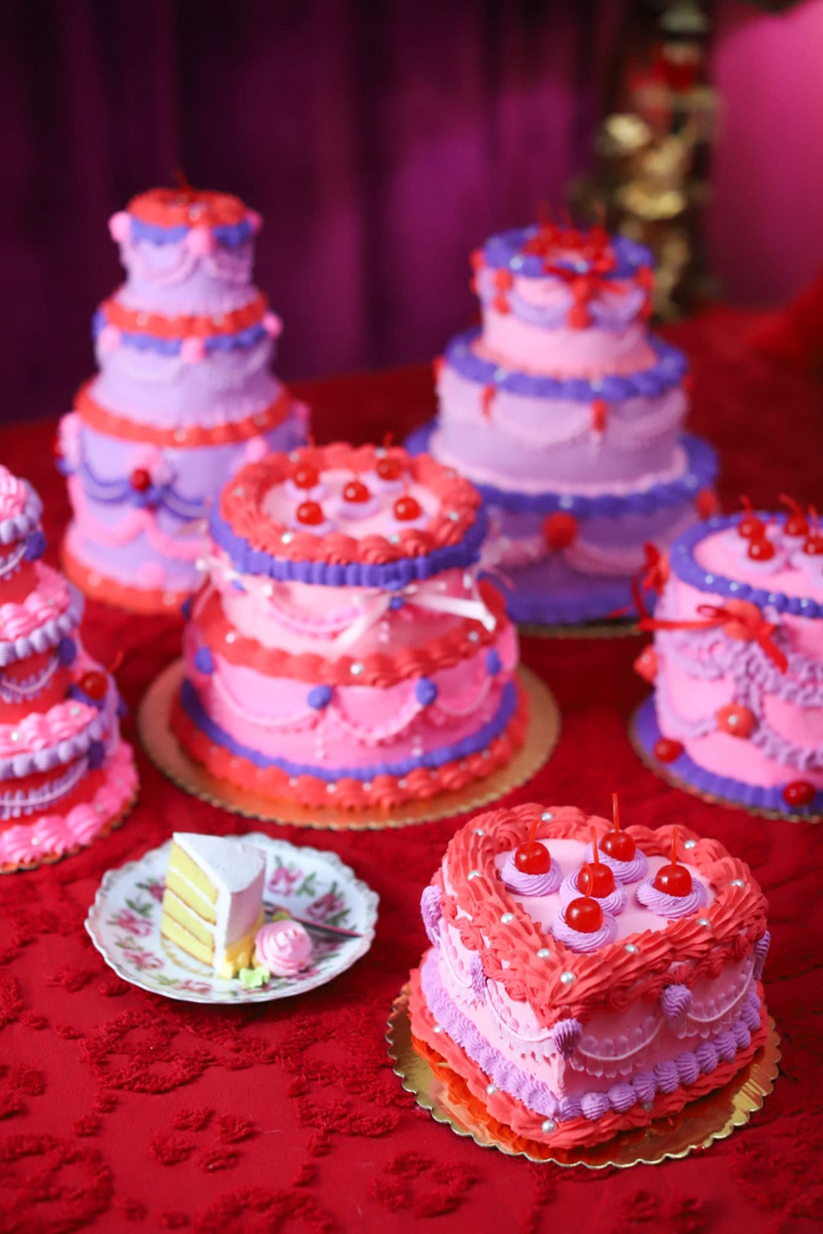 The image shows a variety of colorful and layered cakes arranged on a red tablecloth, with some decorated with cherries and others featuring intricate frosting designs.