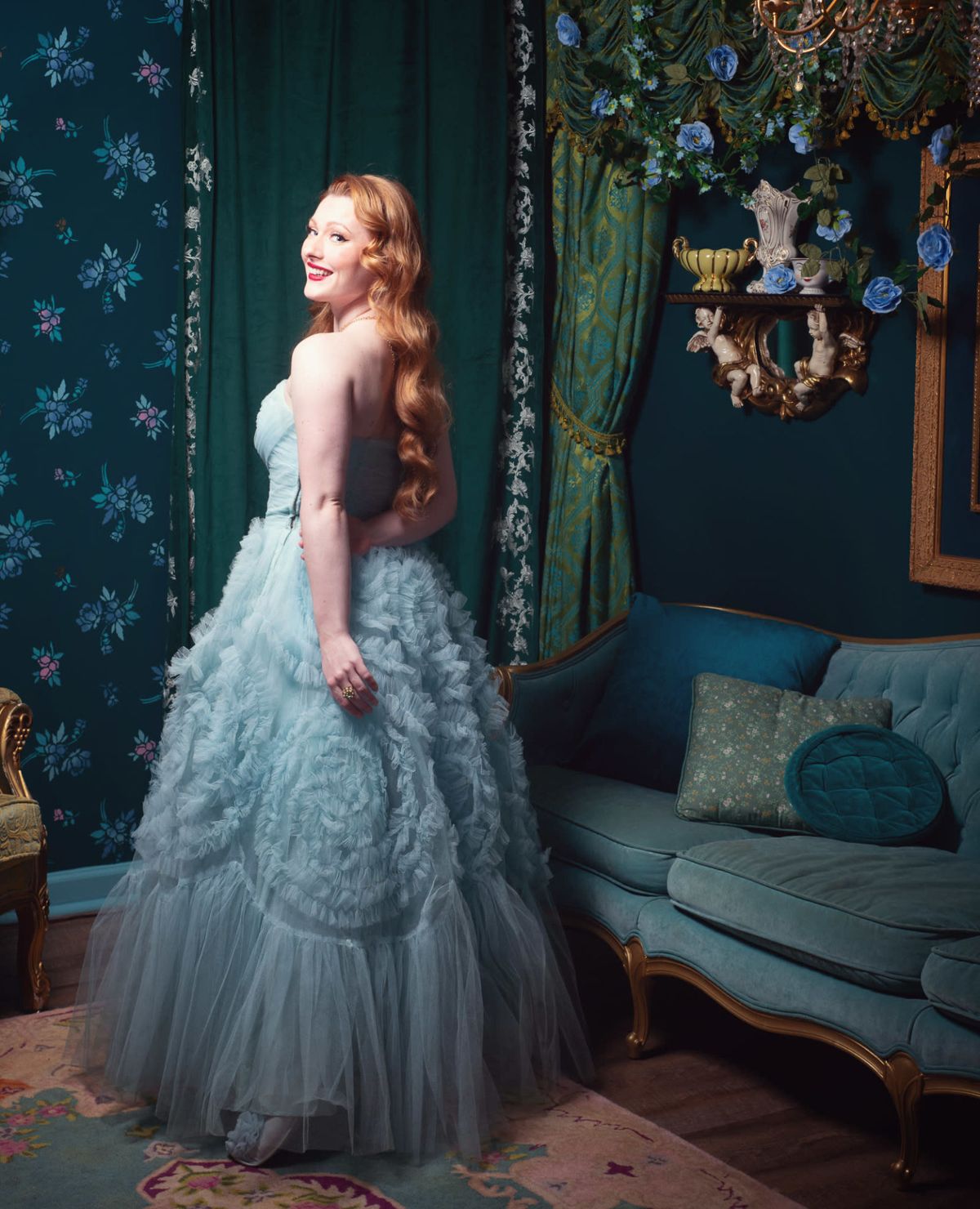 A woman in a light blue, ruffled ball gown stands near a teal couch in a room adorned with floral wallpaper and decorative elements.