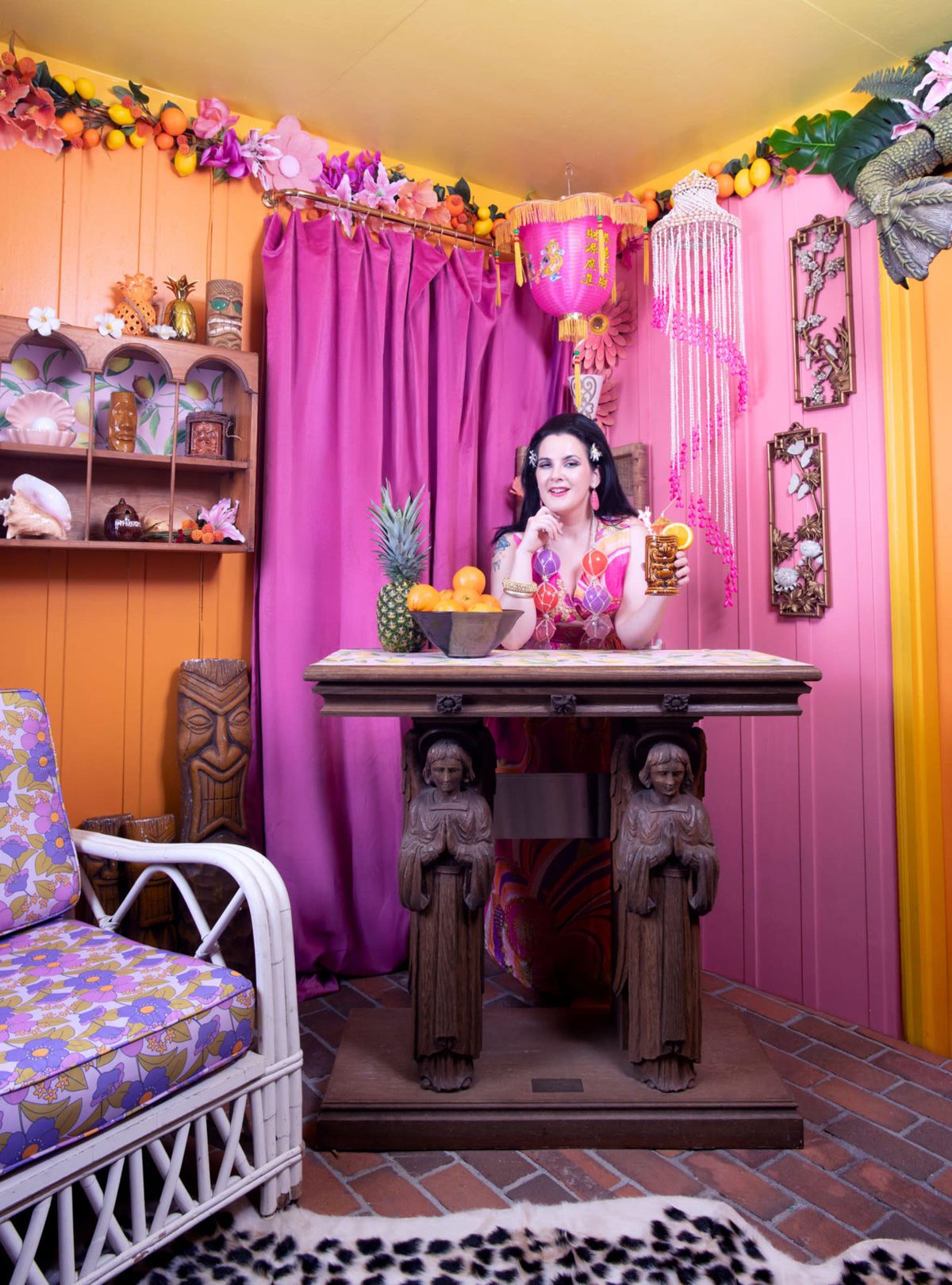 A woman in colorful attire sits at a carved wooden table adorned with tropical fruits in a vibrant, themed interior decorated with curtains and various ornaments.