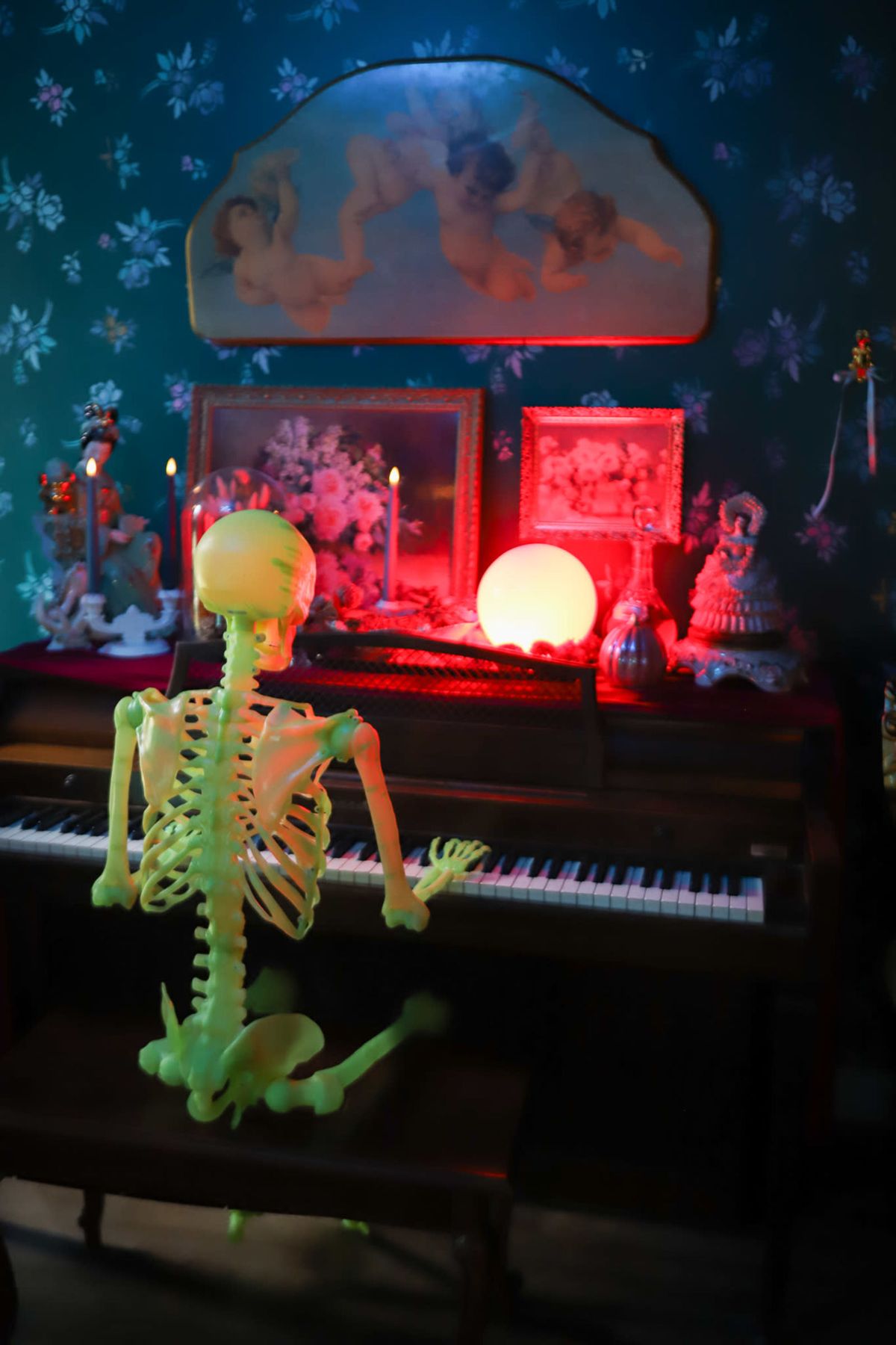 A glow-in-the-dark skeleton sits at a piano surrounded by a vintage decor featuring framed art, candles, and various ornamental items.