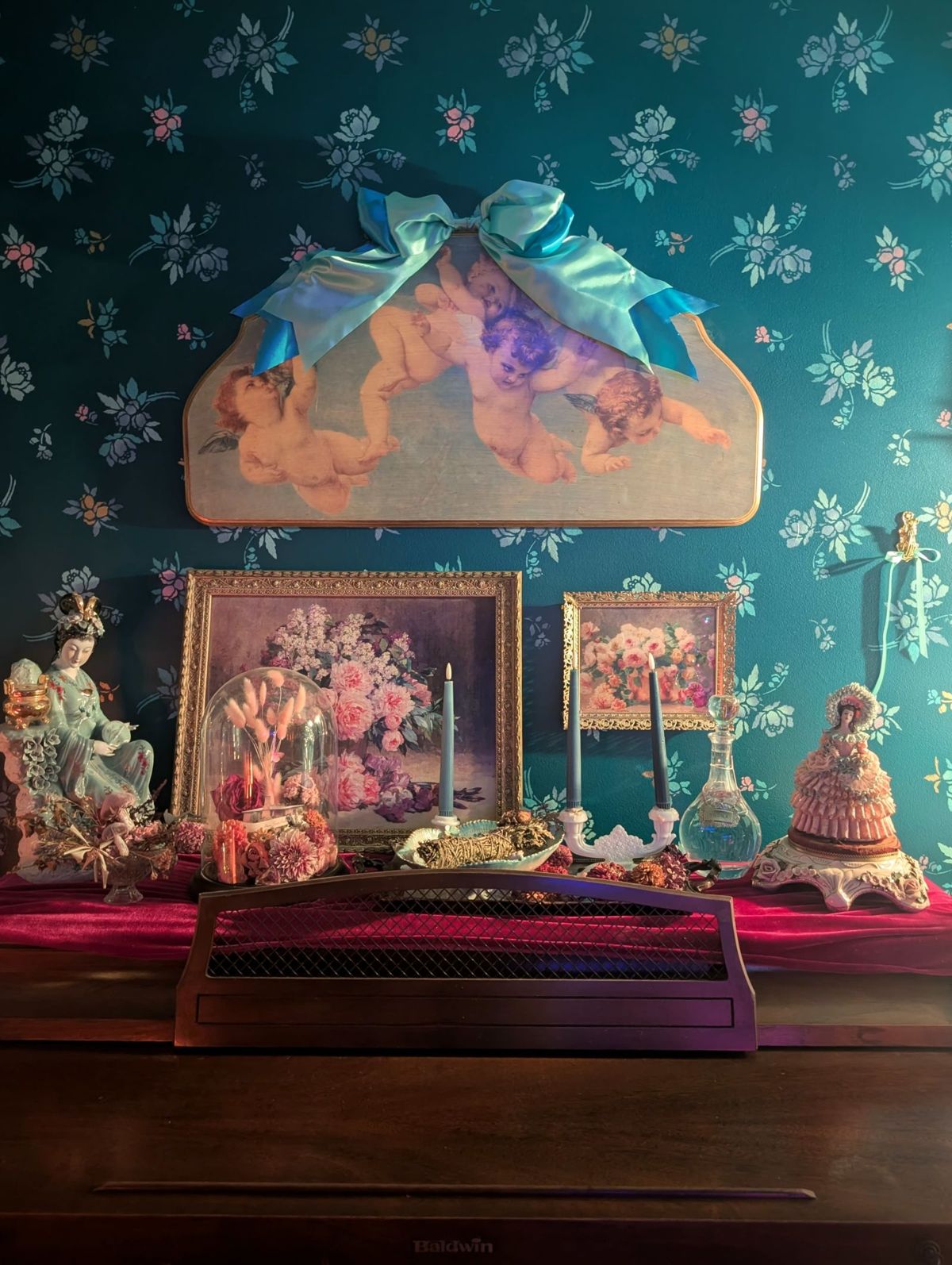 The image features a vintage display with a floral wallpaper backdrop, various framed artworks, figurines, and decorative items arranged on a wooden surface.