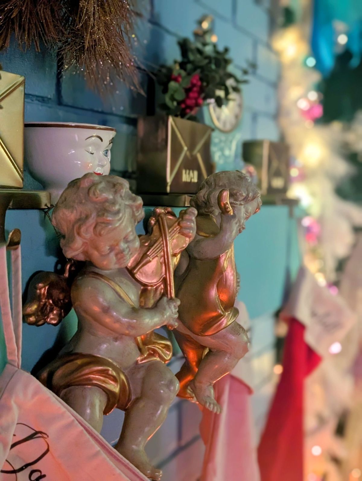 The image features two cherubic figures playing musical instruments, mounted on a blue wall adorned with festive decorations and colorful lights.